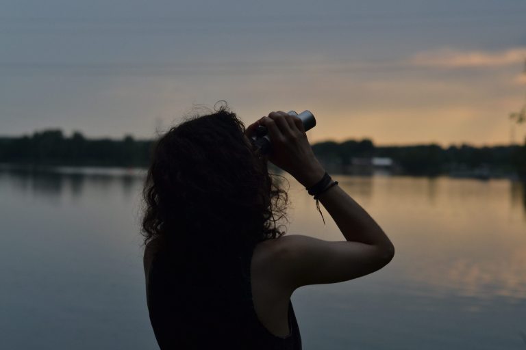 See Vor einem See im Abendlicht steht eine Frau und schaut mit einem Fernglas in den Himmel.