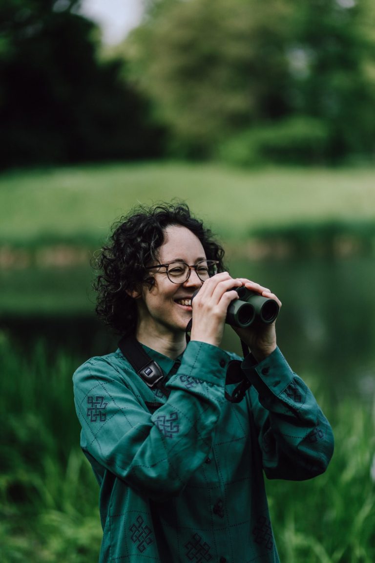 Junge Frau mit Brille und Locken hält ein Fernglas in der Natur.