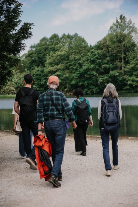 Fünf Personen gehen am Ufer eines Gewässers entlang, umgeben von Bäumen und Natur.